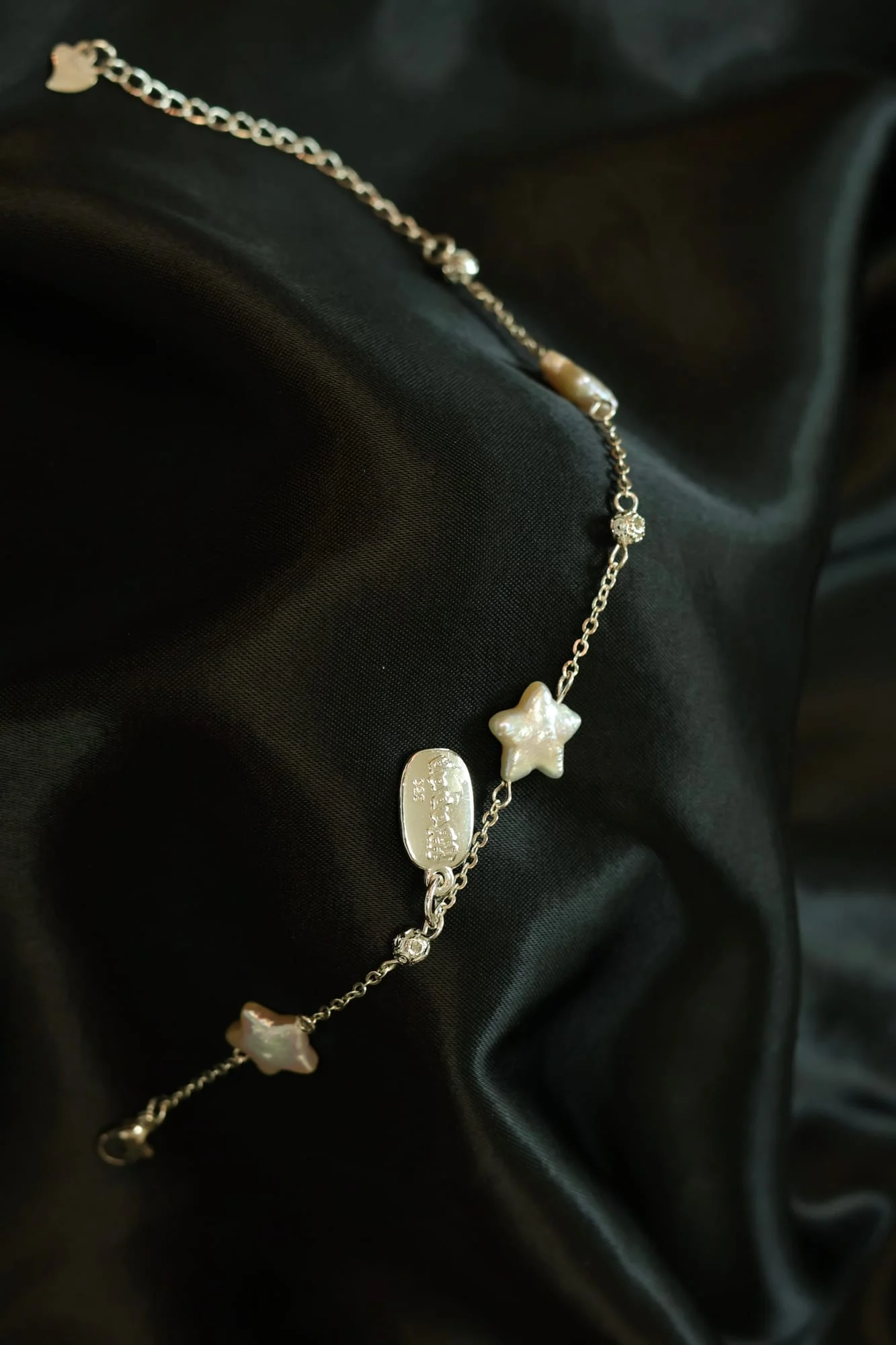 Fine silver-toned chain bracelet with two white iridescent stars, a small oval bead, textured beads and a stamped oval tag, with lobster clasp and extender chain, on black satin.