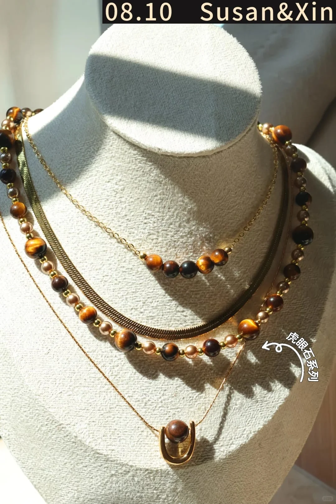 Several gold-toned necklaces layered on a display bust: a tiger’s eye bead strand, a flat chain, a fine chain with five tiger’s eye beads and a single-bead arc pendant.